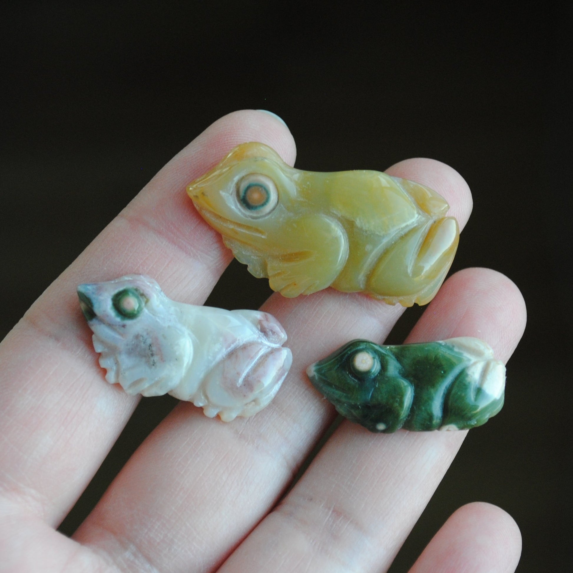 Three small stone frog carvings held in a hand against a blurred background