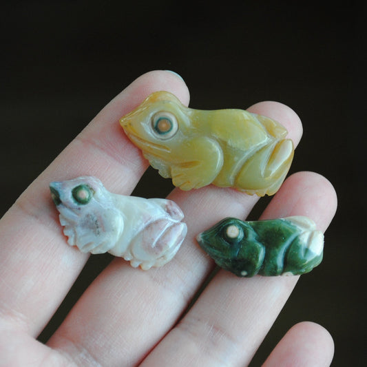 Three small stone frog carvings held in a hand against a blurred background