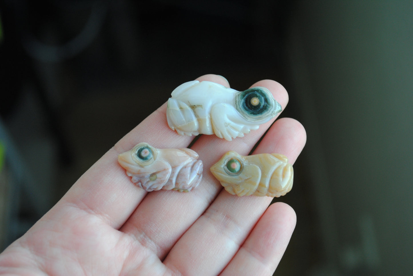 Three small stone frog carvings held in a hand against a blurred background