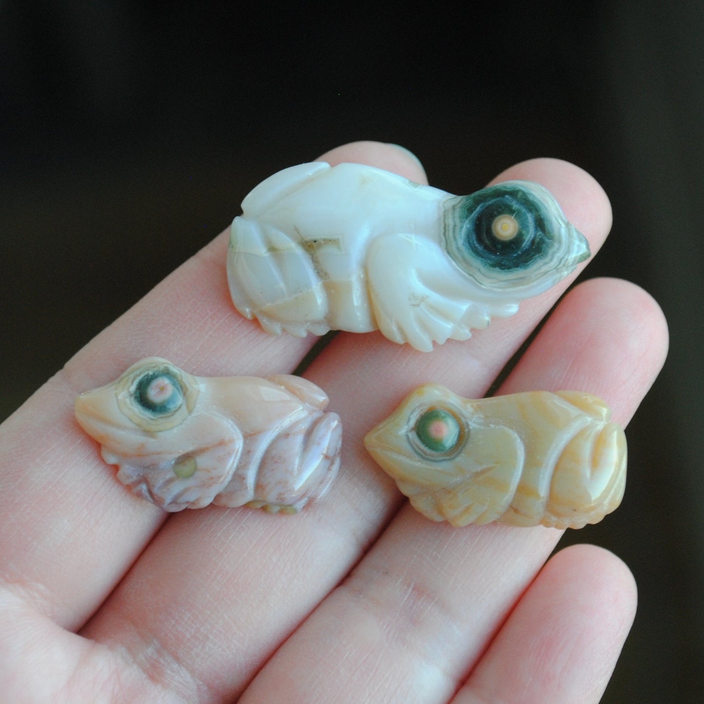 Three small stone frog carvings held in a hand against a blurred background