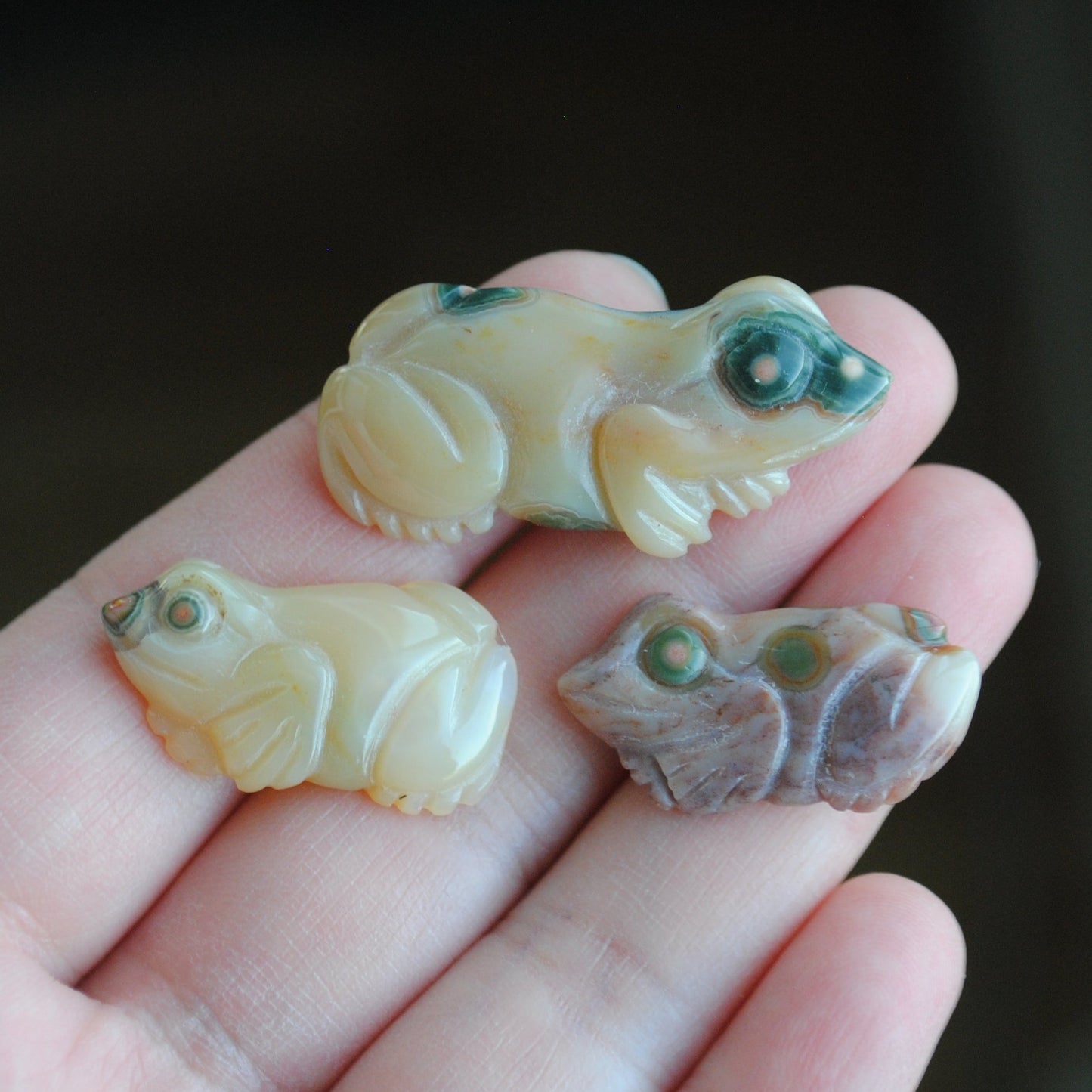 Three small stone frog carvings held in a hand against a blurred background
