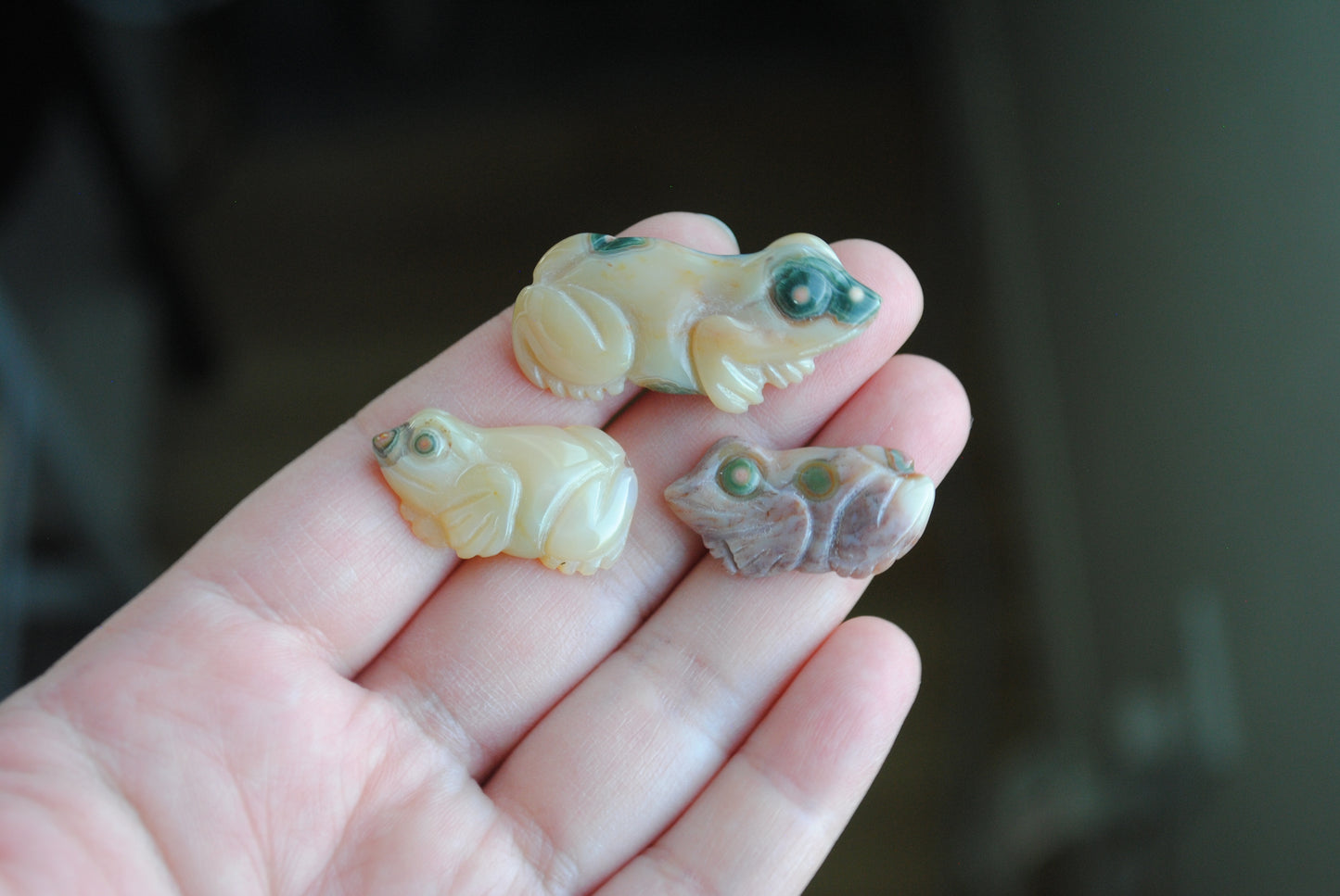 Three small stone frog carvings held in a hand against a blurred background