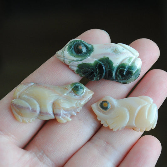 Three small stone frog carvings held in a hand against a blurred background