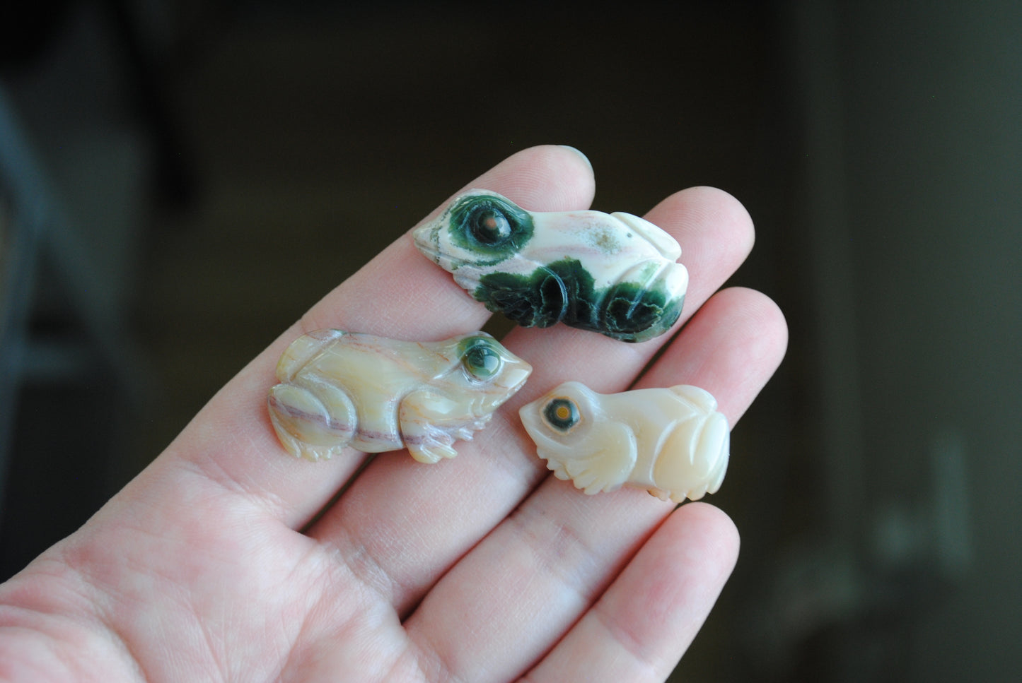 Three small stone frog carvings held in a hand against a blurred background